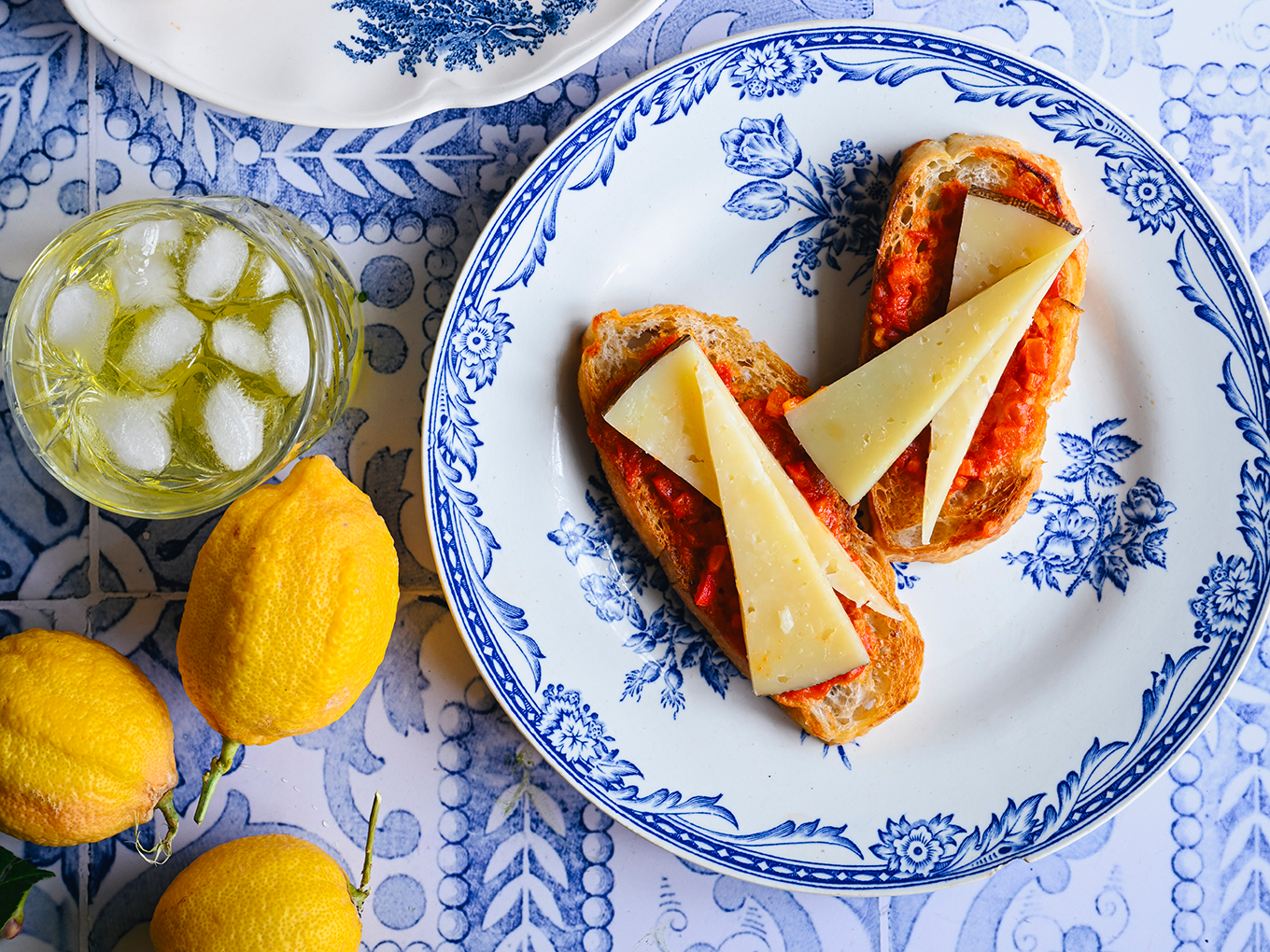 Paahdettuja tomaattileipiä eli pan con tomate manchegojuustolla sinivalkoisilla lautasilla sitruunoiden ja juomien vieressä