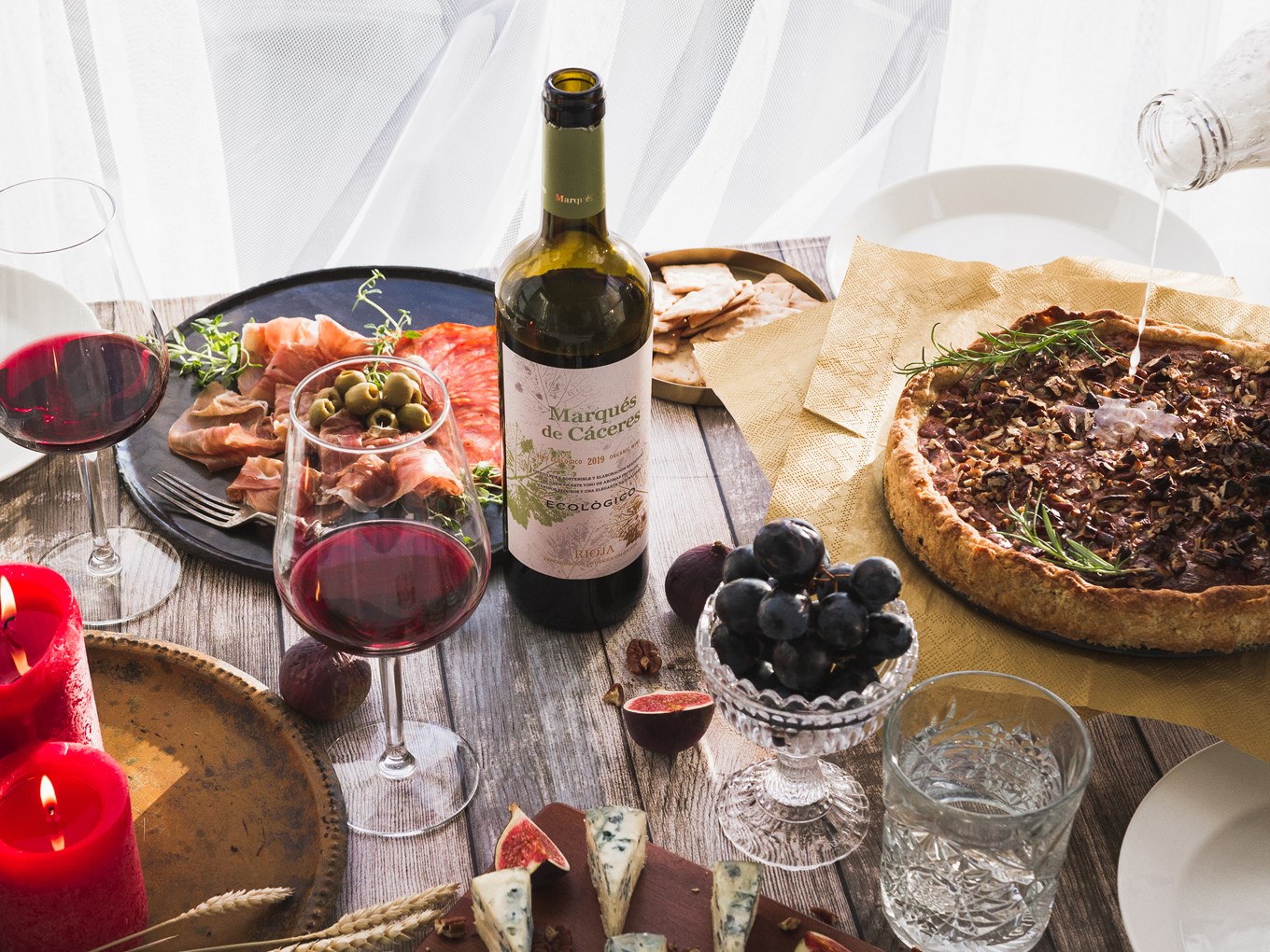 Tunnelmallinen illallisistujaisten tapaspöytä, jossa Manchego-pekaanipähkinäpiirakka, juustoja, leikkeleitä, hedelmiä sekä pari lasia Marqués de Cáceres -punaviiniä